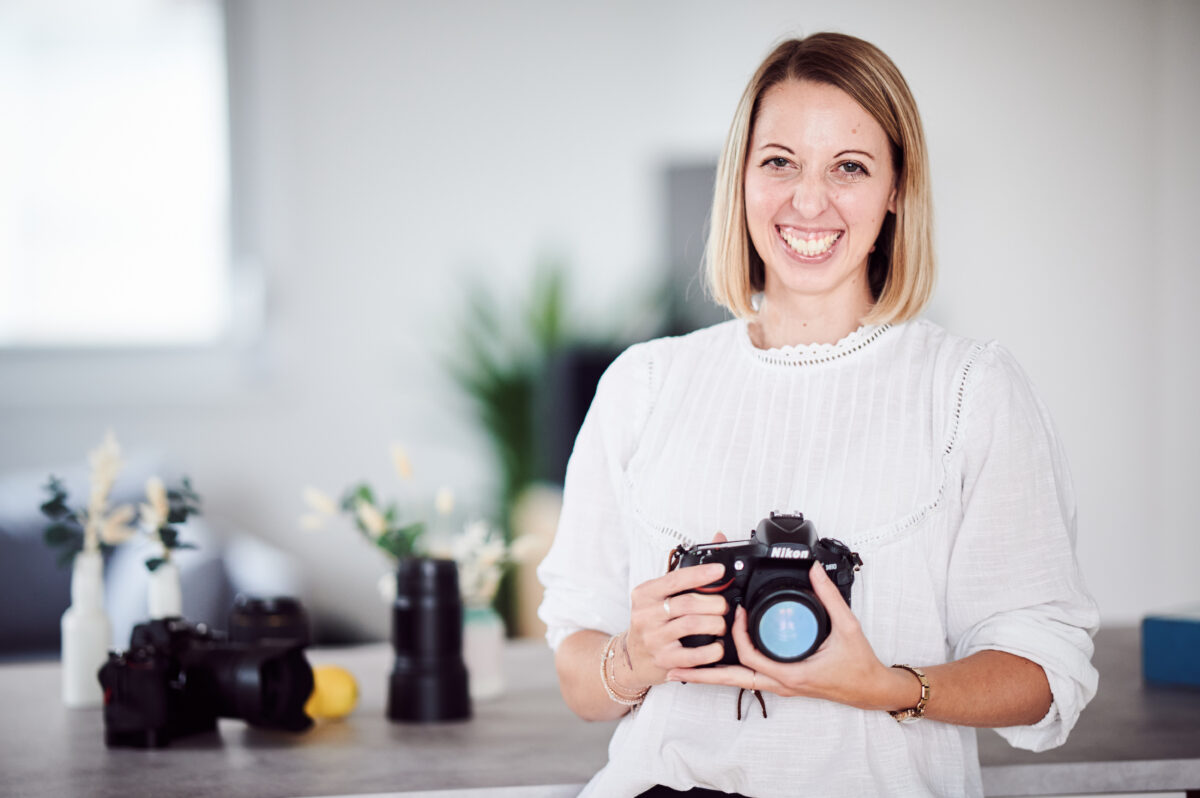Food Fotografin Cornelia hält eine Spiegelreflexkamera in ihren Händen und lächelt in die Kamera