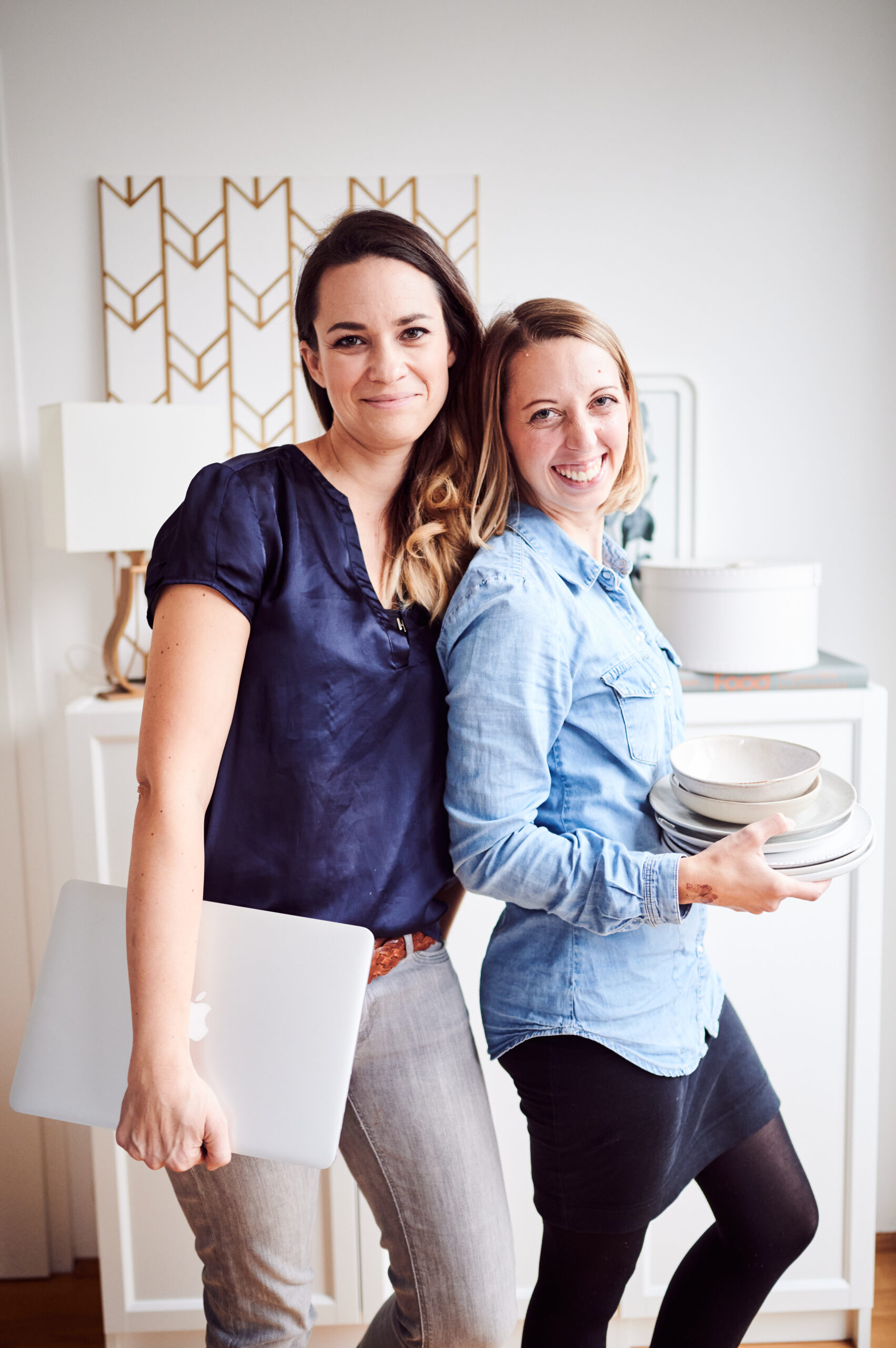 Mariella & Cornelia vom Food Foto Campus vor einem weißen Schrank mit Tellern und Laptop in der Hand